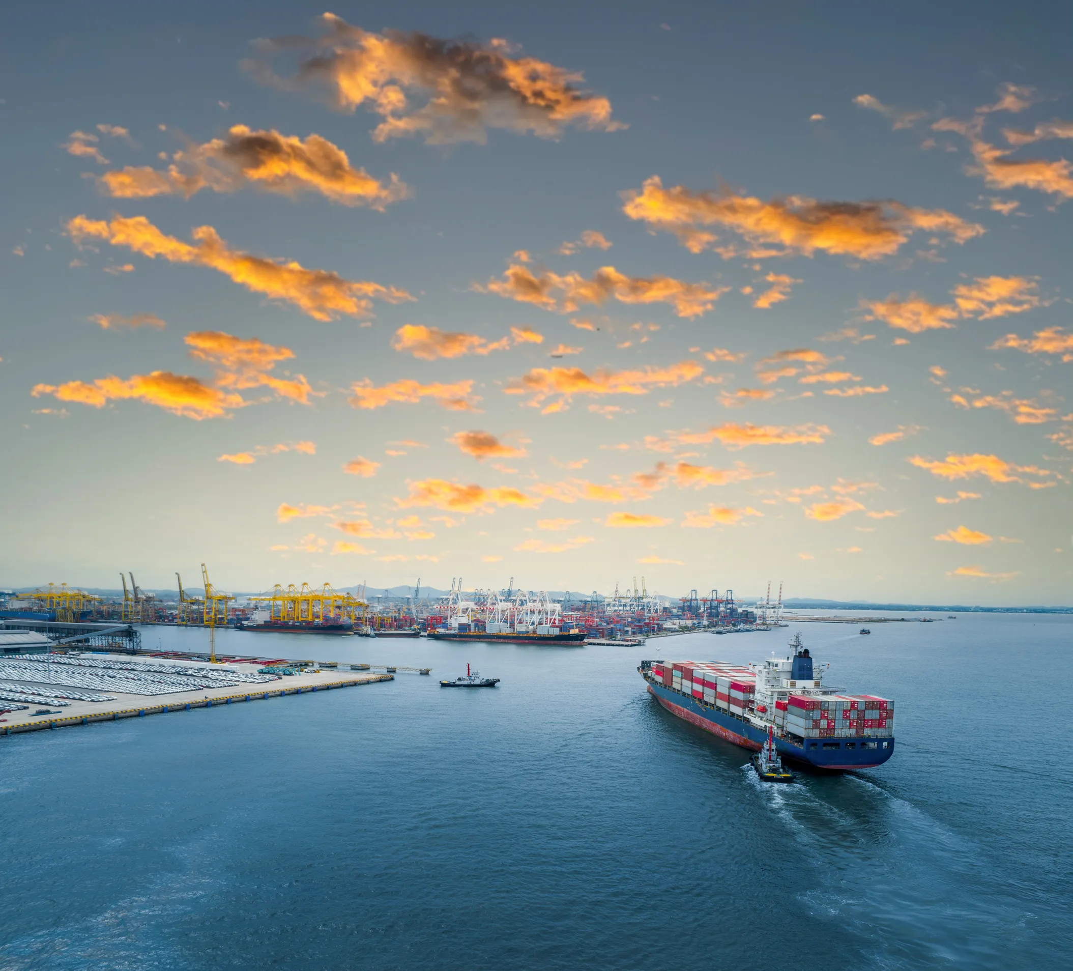 Puerto marítimo con grúas de carga y un buque portacontenedores saliendo al mar al atardecer